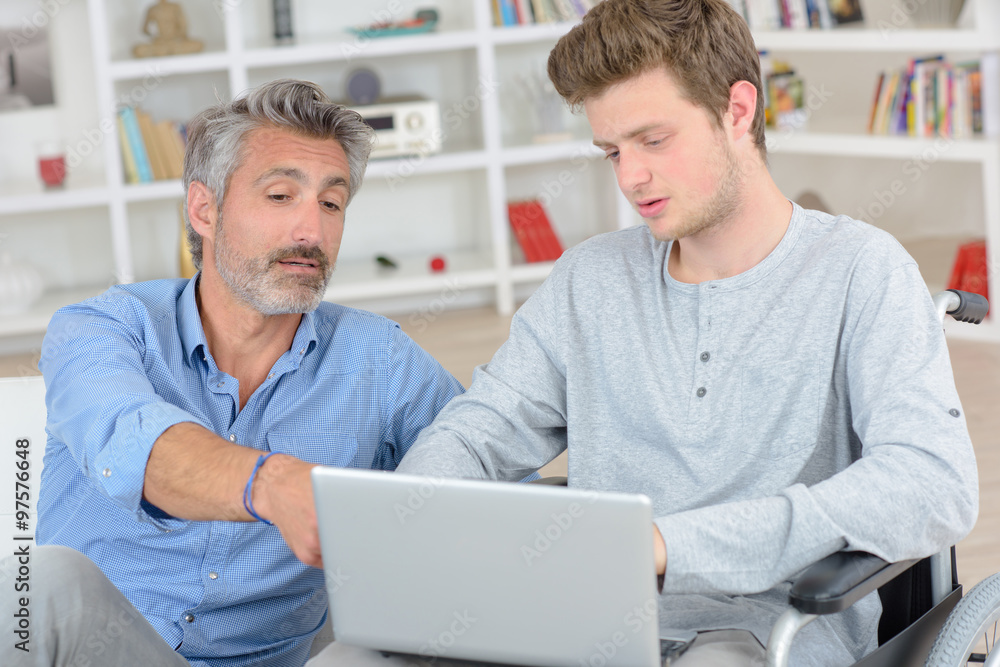 Fototapeta premium Father pointing to screen of laptop held by son in wheelchair