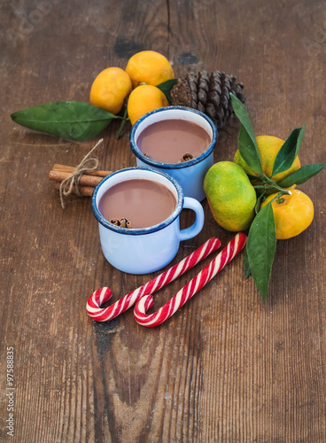 Hot chocolate in enamel metal mugs, fresh mandarins, cinnamon sticks, pine cone and candy canes over rustic wooden background