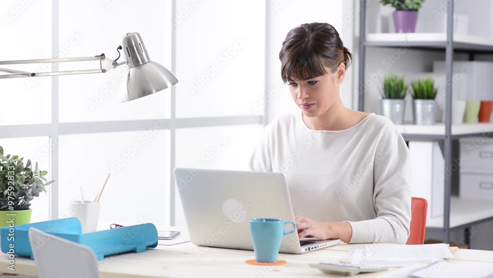 Smiling young secretary working at office desk, typing on a laptop and looking at camera Smiling young secretary working at office desk, typing on a laptop and looking at camera