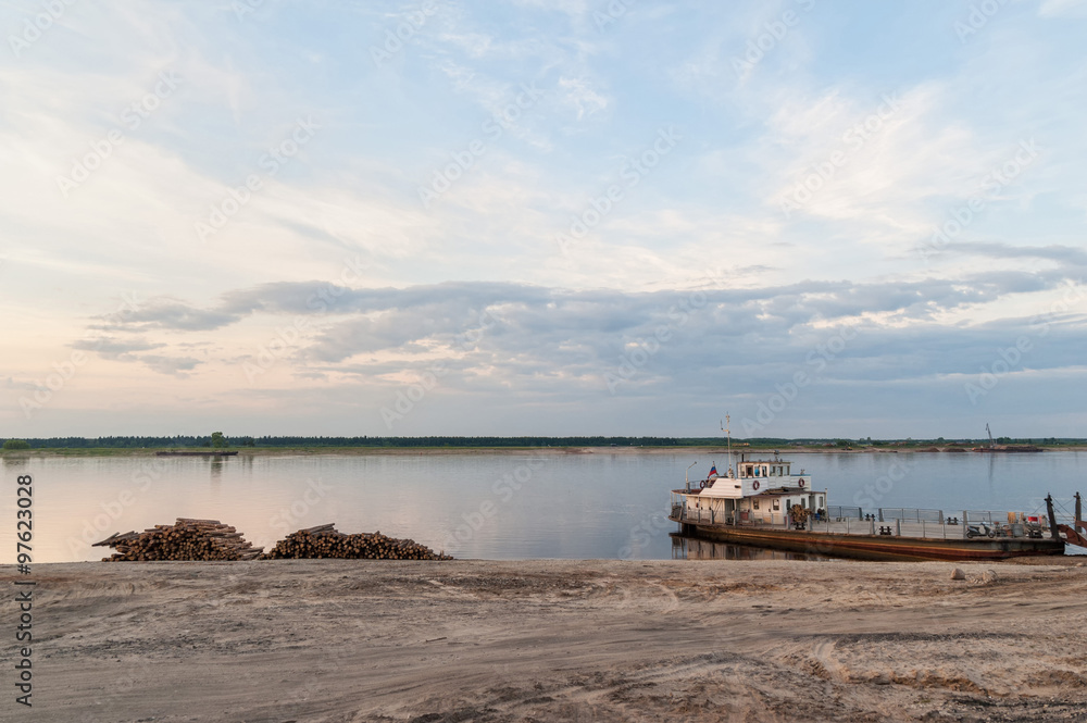 Timber store sinks in high water along riverside, ferry and cargo ship against cloudy sky background at sunset. Severnaya Dvina River, Bereznik settlement, Arkangelsky region, Russia.
