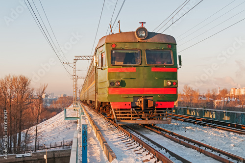 Green suburban electric train moves towards on snow-bound railroad vanishing in horizon before sunset light against skyline background. Moscow, Russia. 