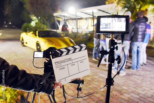 Operator holding clapperboard during the production of short film outdoor in the night with sportive yellow car and actor on stage. Focus on the clapperboard and monitors photo