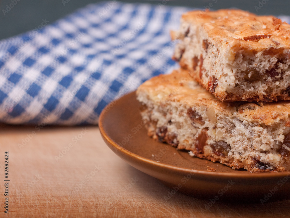 Closeup view of two piece of biscuit cake with walnuts and raisins on brown ceramic saucer, with blue plaid cloth on the background. Closeup view of two piece of biscuit cake with walnuts and raisins on brown ceramic saucer, with blue plaid cloth on the background.