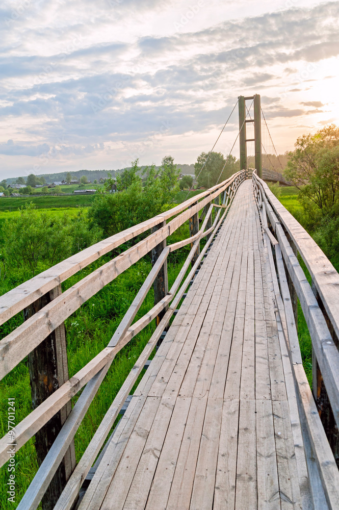 Old lengthy hanging wooden footbridge with rails over river against sunset background. Arkhangelsky region, Russia. 
