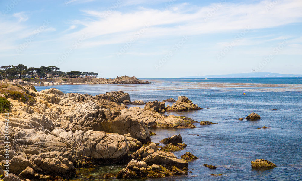 Rocky shoreline in Monterey Bay with a crowded beach, boats in the background and a kayaker.