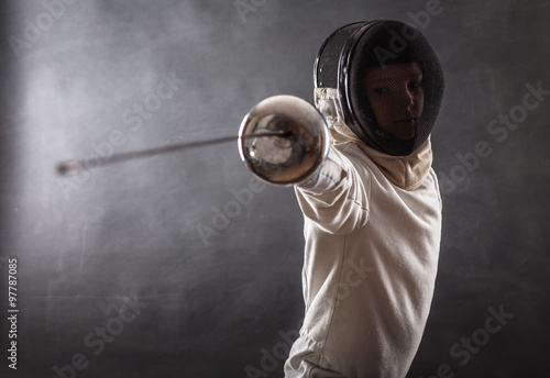 Boy wearing white fencing costume and black fencing mask standing with the sword practicing in fencing Fototapeta