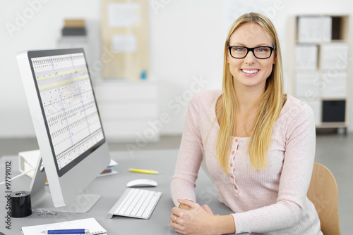 Fotografie motivierte mitarbeiterin sitzt am schreibtisch im büro mit tabellen auf dem moni