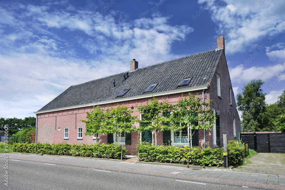 Renovated brick farmhouse with trees in front, cloudy blue sky, Netherlands.