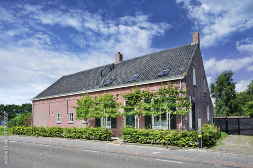 Fototapeta Naklejka Na Ścianę i Meble -  Renovated brick farmhouse with trees in front, cloudy blue sky, Netherlands.