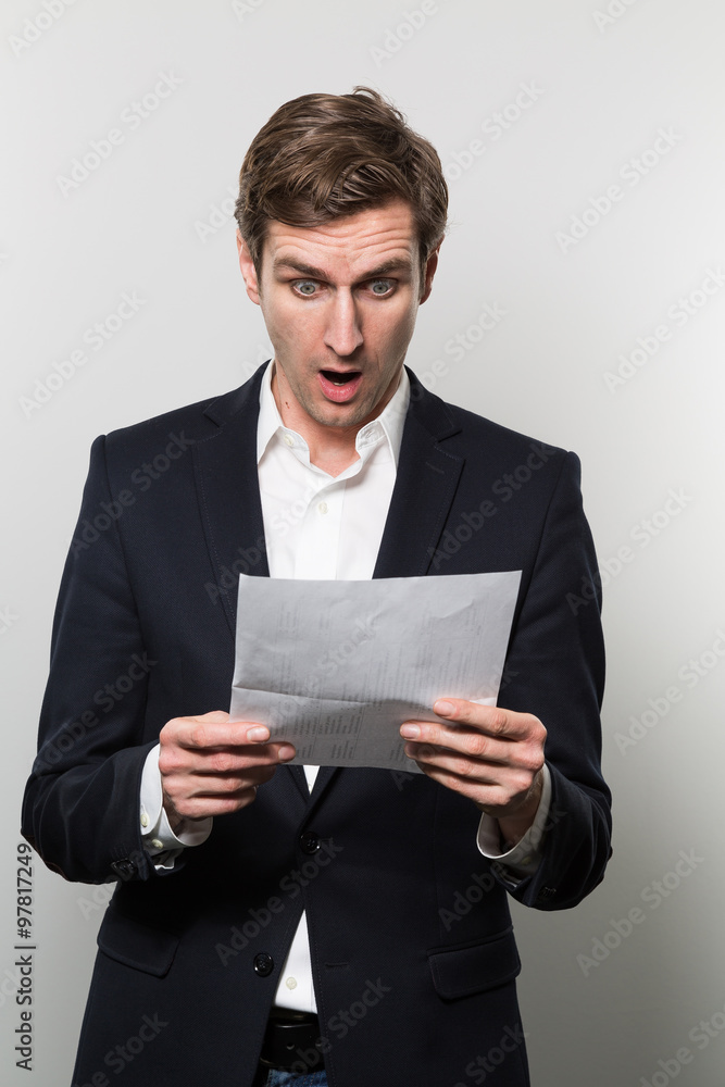 Blond-haired european businessman with a scared look stares at a piece of paper with bad news on it while in front of a gradient background Blond-haired european businessman with a scared look stares at a piece of paper with bad news on it while in front of a gradient background