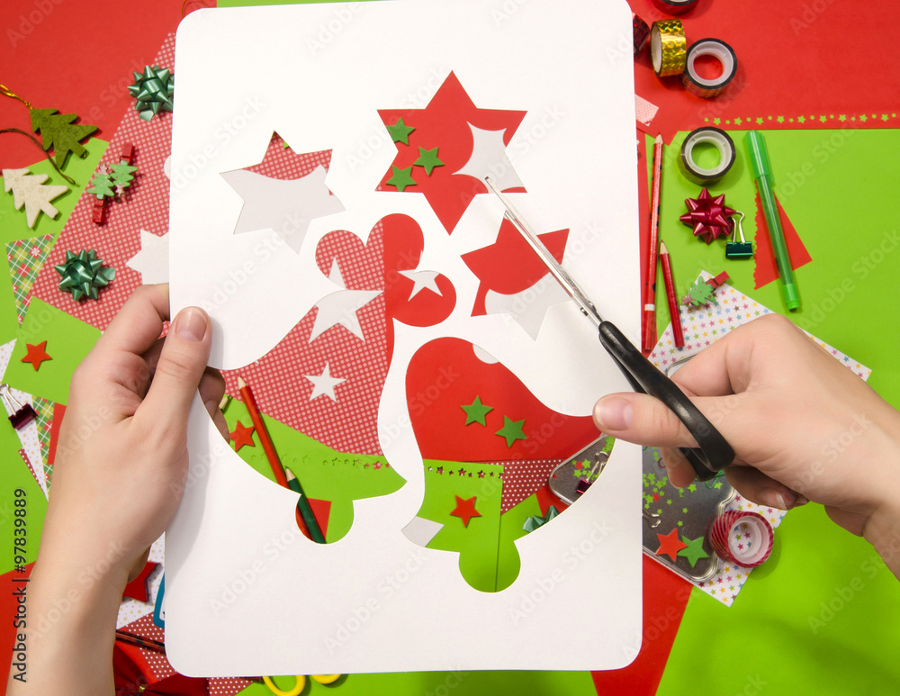Fototapeta premium Hand of a woman cutting a white cardboard with Christmas shapes,stars and bells.Red and green paper,pencils,craft scissors, cardboard bell cut and decorations.Arts and craft supplies for Christmas.