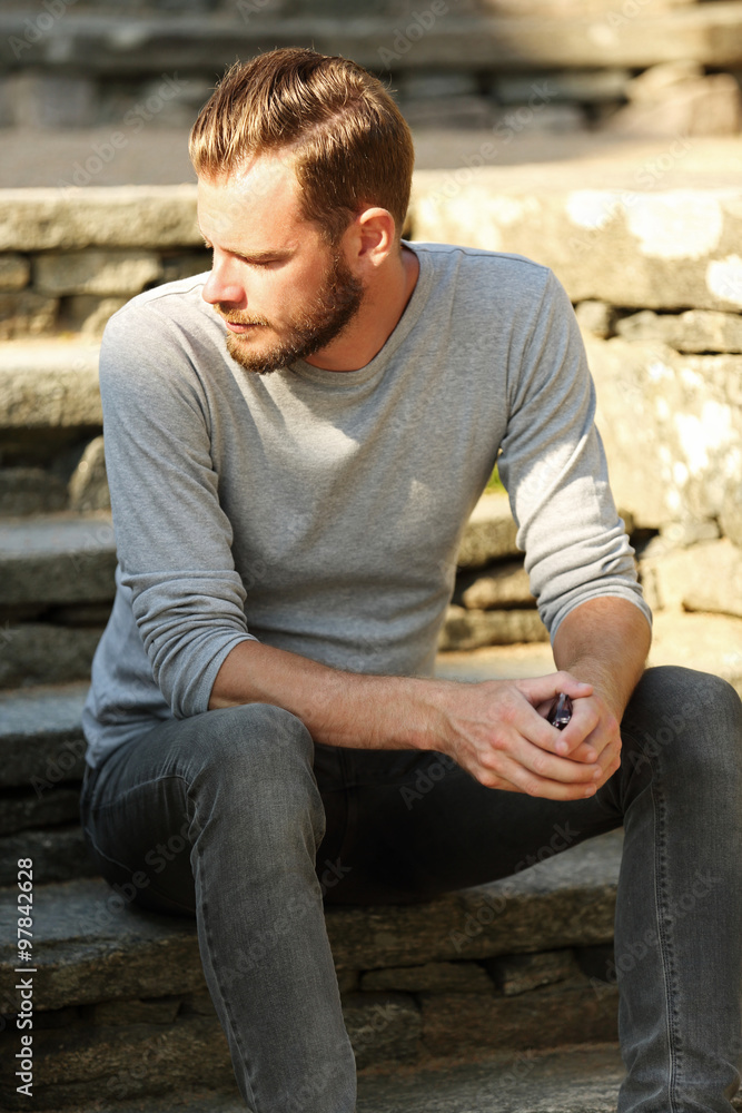 Man in his 20s wearing a grey shirt and jeans, sitting down outside on a set of steps on a sunny summer day. Man in his 20s wearing a grey shirt and jeans, sitting down outside on a set of steps on a sunny summer day.