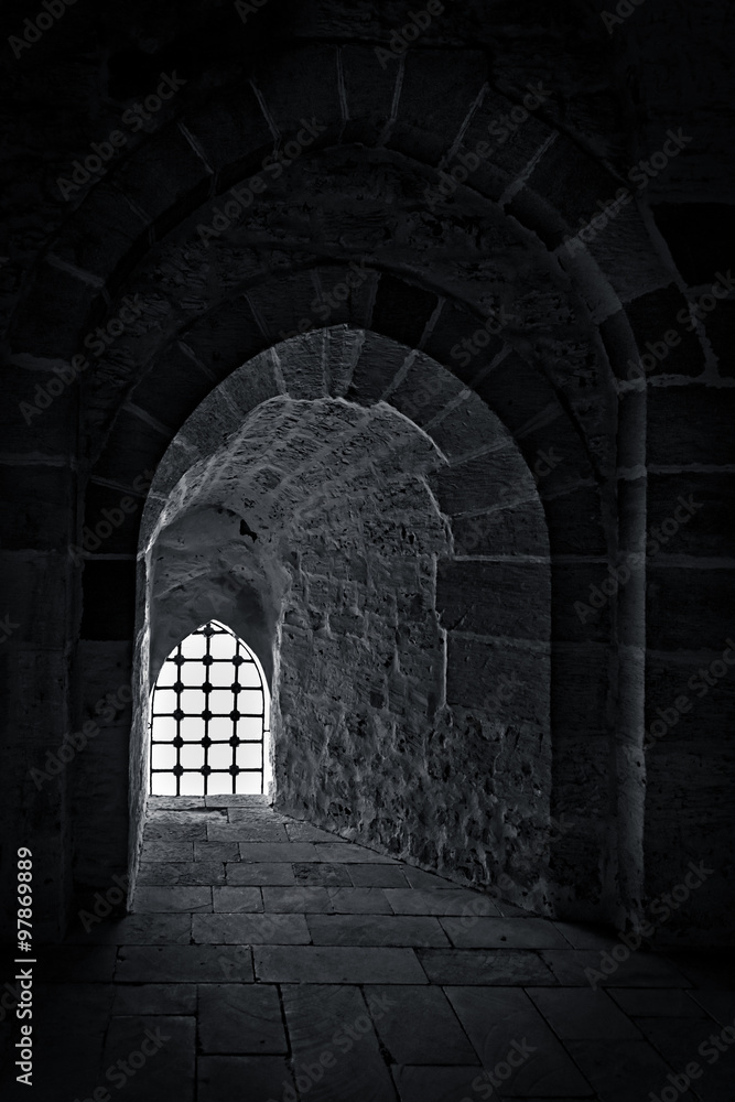 Fototapeta premium Stone wall with one backlit window with iron grid at one of the passages of an old citadel, Alexandria, Egypt. A fifteenth century defensive fortress located on the Mediterranean sea coast