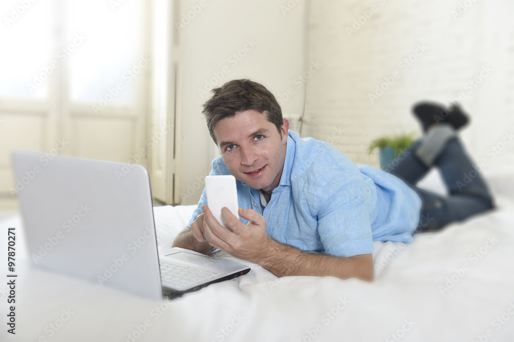 young attractive man lying on bed using mobile phone and computer laptop working from home young attractive man lying on bed using mobile phone and computer laptop working from home