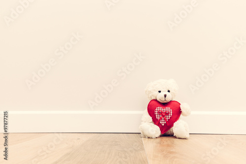 Cute teddy bear with big red plush heart. Sitting on wooden floor against white wall.