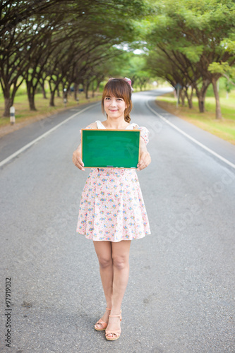 Cute woman hand holding green blank board sign on road with tree, Smiling female model.