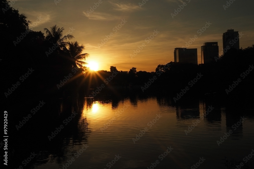 Sunrise over pond at park in a city. Silhouette tree and building against with sunrise, orange sky and sun ray.