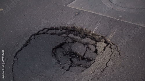 Handheld camera goes up to a dangerously large sinkhole near a manhole cover in an asphalt topped road and then pulls the shot away from the pothole. photo