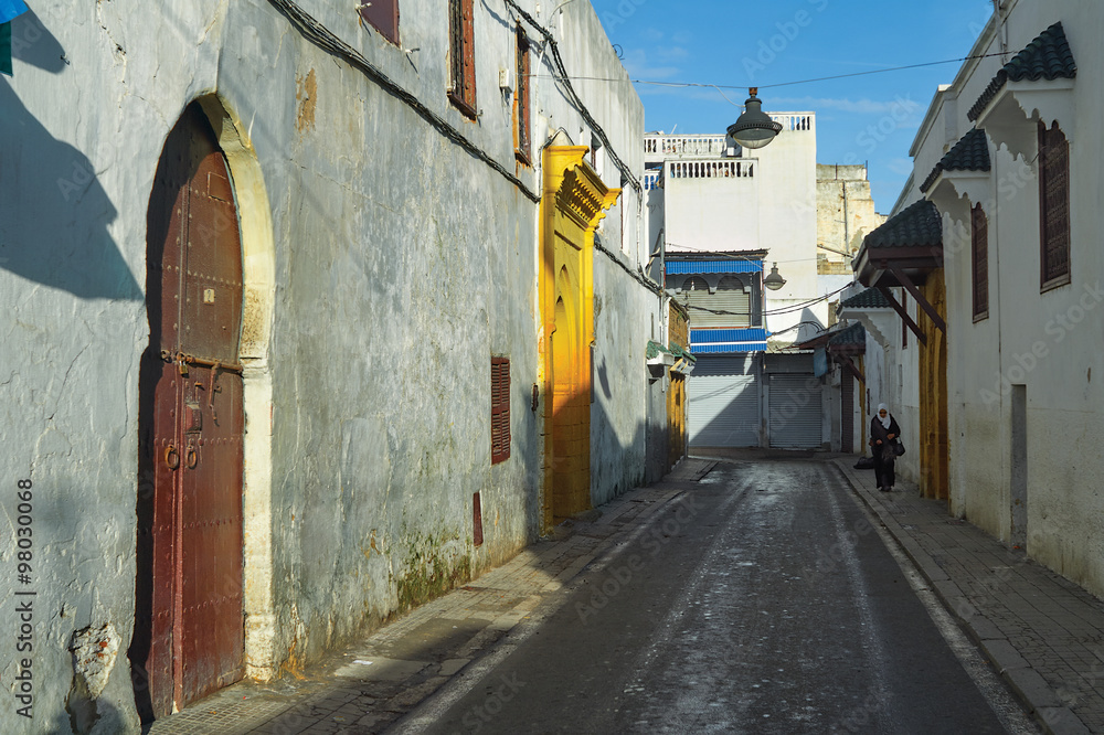 Obraz premium The walls of the ancient city and a woman, dressed in the Arab national clothes, walking through the old streets of the town of Sali (Morocco) on a sunny spring day