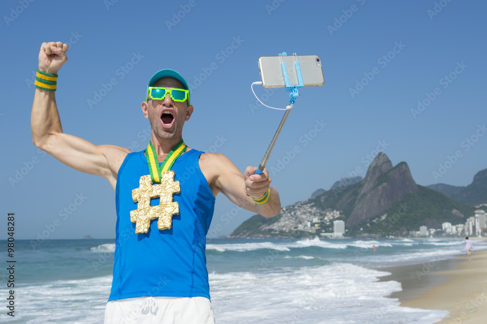 Hashtag gold medal athlete punching the air as he poses for a picture with  his mobile phone on a selfie stick on Ipanema Beach in Rio de Janeiro,  Brazil Stock Photo