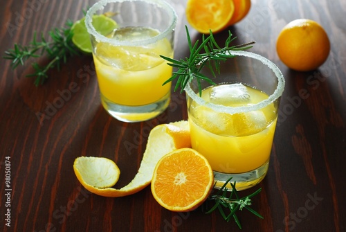 Two sugar-rimmed glasses of tangerine winter cocktails with ice, with tangerines, tangerine peel, lime and rosemary on brown wooden background. Selective focus.