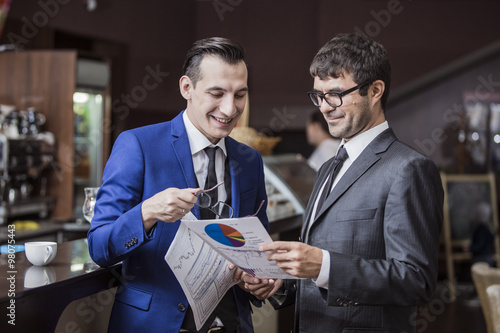 Два молодых бизнесмена на переговорах в кафе изучают документы и радуются успехам.