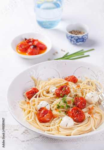 .Italian spaghetti pasta with baked cherry tomatoes, mozzarella and spring onions in a white bowl on a white table.