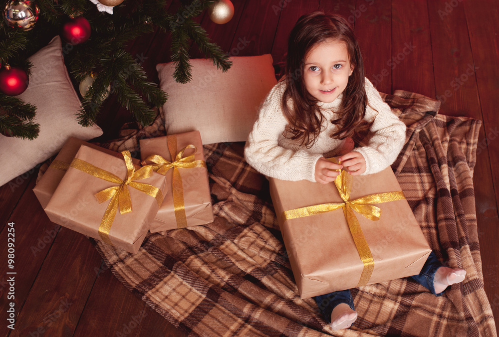 Cute kid girl 4-5 year old opening christmas presents in room. Wearing trendy knitted sweater. Sitting on wooden floor. Looking at camera. Cute kid girl 4-5 year old opening christmas presents in room. Wearing trendy knitted sweater. Sitting on wooden floor. Looking at camera.