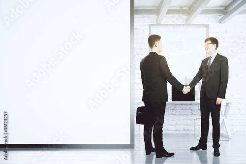 Businessmen shake hands in loft office with blank white wall, mo