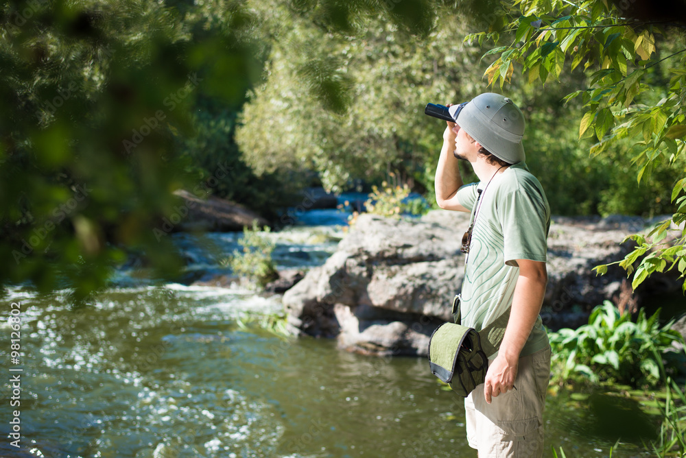 Man in pith helmet looking up through binoculars by river Man in pith helmet looking up through binoculars by river