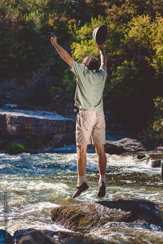 Man holding binoculars jumping up with pithhelmet on rocky riverbank photo