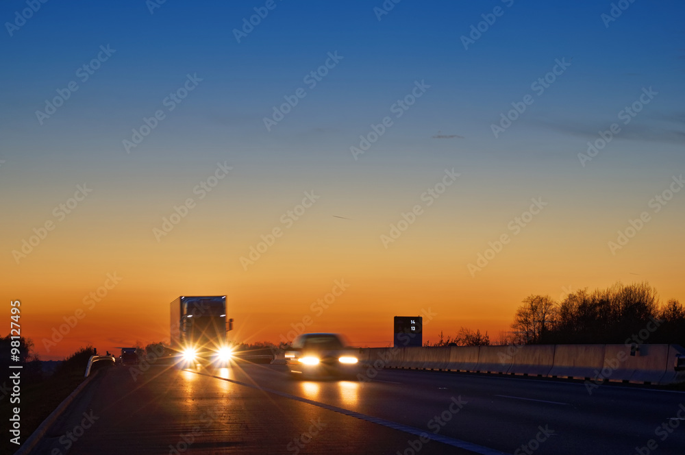 Highway with oncoming trucks and a car after sunset. Shining the spotlight cars. Information boards about the temperature. Blue and orange sky is clear.