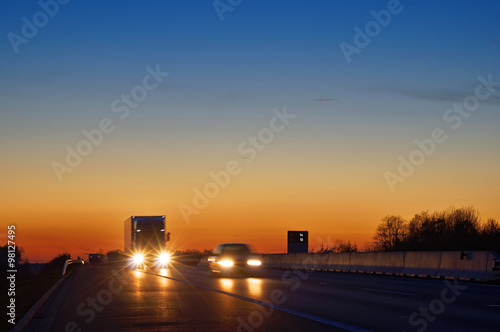 Obraz na płótnie Highway with oncoming trucks and a car after sunset. Shining the spotlight cars. Information boards about the temperature. Blue and orange sky is clear. w salonie