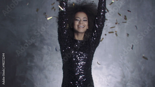 Sexy afro american woman dancing among confetti over silver background. Beautiful black woman throwing golden confetti, slow motion .