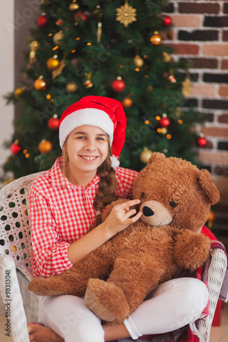 holidays, presents, christmas, childhood and people concept - smiling girl in santa helper hat with teddy bear over lights background