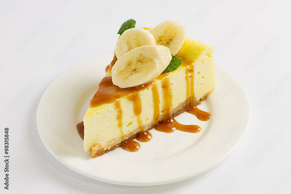 Slice of cheesecake topped with caramel sauce, banana and mint on plate on white table, close up Slice of cheesecake topped with caramel sauce, banana and mint on plate on white table, close up