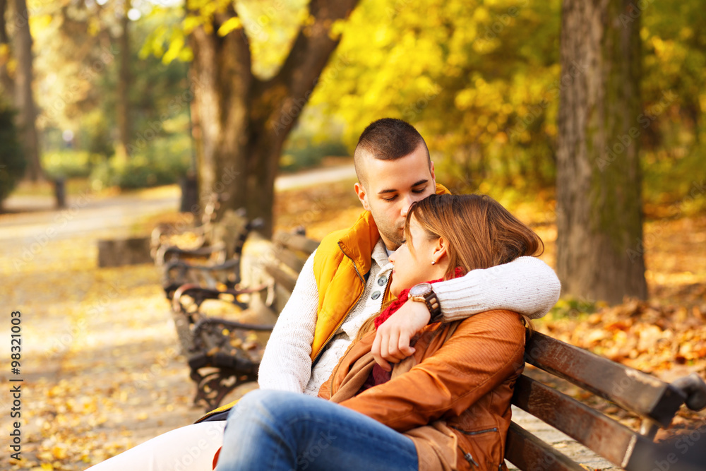 Young couple sitting on a park bench on a sunny autumn day and expressing tenderness Young couple sitting on a park bench on a sunny autumn day and expressing tenderness