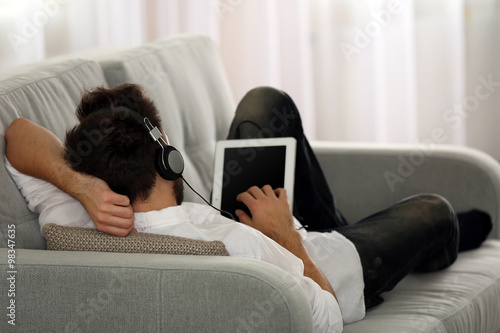 Young man listens music with headphones on grey sofa in the room