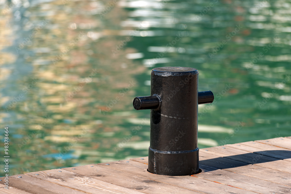 Black Mooring Bollard / Detail of old and black mooring bollard on the wooden pier with water in the background Black Mooring Bollard / Detail of old and black mooring bollard on the wooden pier with water in the background