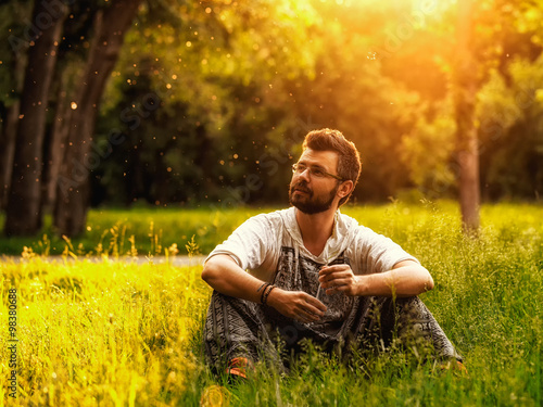 A bearded man sitting on a grass in the park on summer day, looks lost in thought and inspired