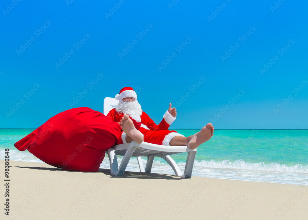 Obraz premium Santa Claus with Christmas sack on deckchair at ocean beach
