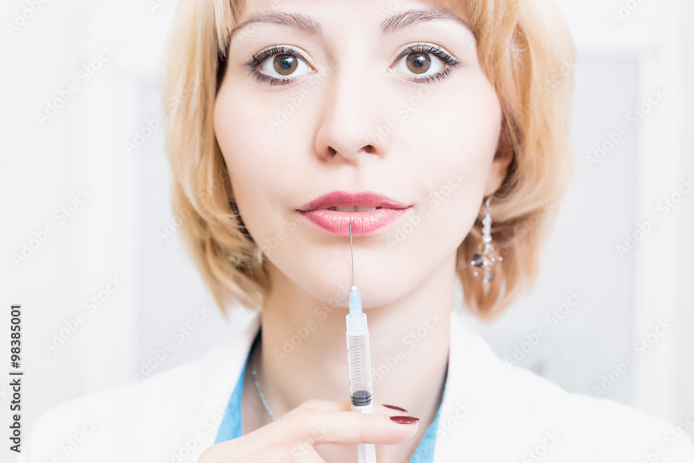 woman beautician is permanent in a white robe in the office and keeps the needle of the syringe near the lips. Doctor with syringe woman beautician is permanent in a white robe in the office and keeps the needle of the syringe near the lips. Doctor with syringe