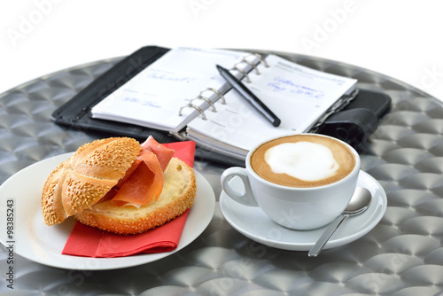 Kaffeepause mit belegtem Sesambrötchen und einer Tasse Cappuccino auf einem Cafeteria-Tisch photo