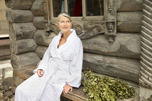 Smiling senior woman in bathrobe sitting outside Finnish sauna log cabin
