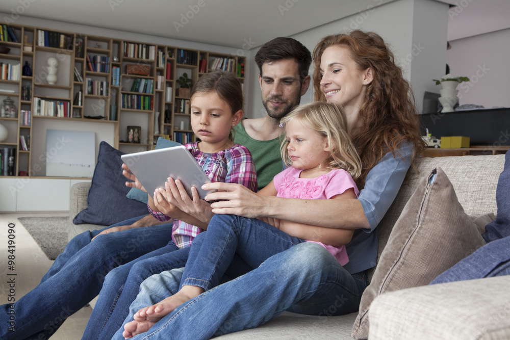 Couple sitting with their with two little daughters on couch in the living room looking at digital tablet Couple sitting with their with two little daughters on couch in the living room looking at digital tablet