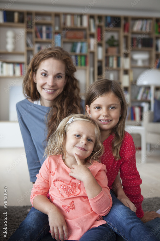Portrait of two little sisters and her mother in the background sitting on the floor at living room Portrait of two little sisters and her mother in the background sitting on the floor at living room