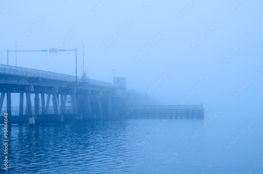 A draw bridge with stop light, gate, bridge keeper's house and channel bumpers is disappearing into a heavy fog.
