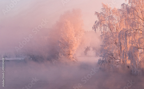 Winter dawn over the misty river, frozen trees illuminated by the red rising sun © alexugalek