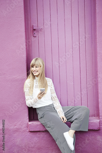 Happy young woman with cell phone sitting by pink doorway