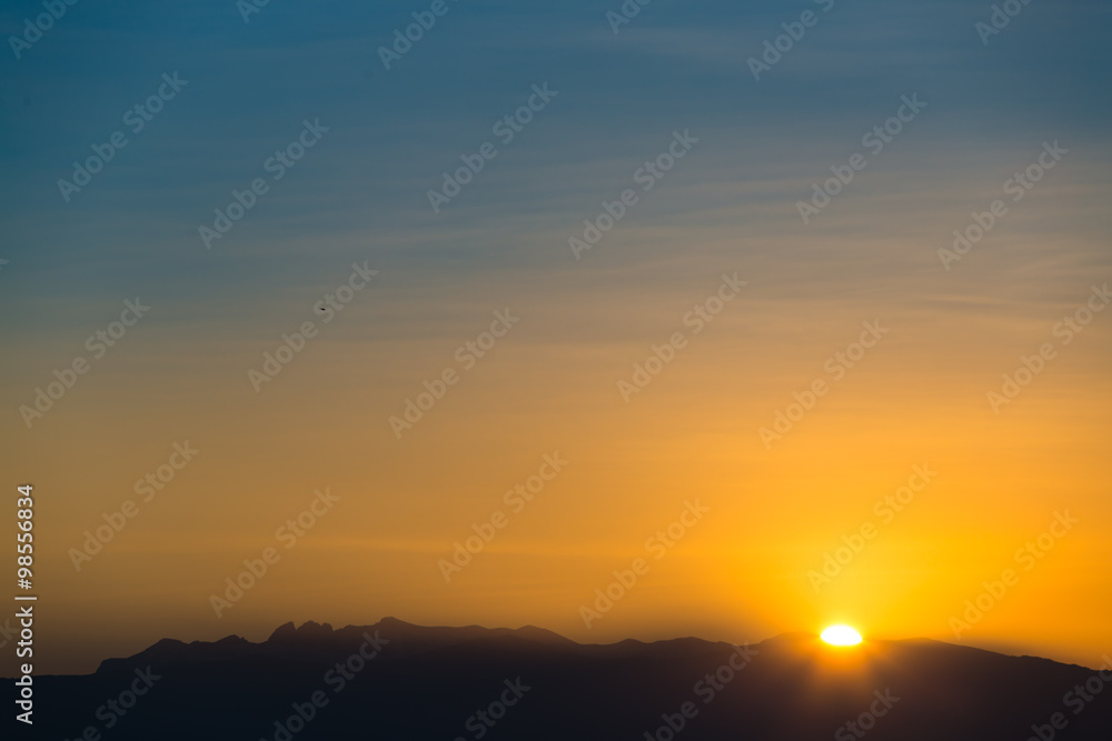 Sun behind dark mountain silhouettes, with blue and orange colored sky